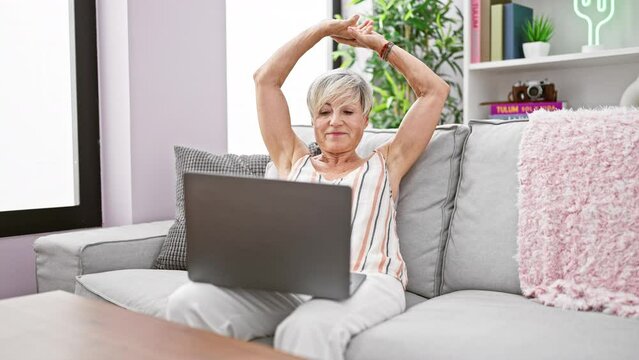 Mature woman using laptop and stretching on sofa indoors