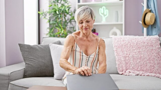 A mature woman with short grey hair relaxes on her living room sofa while closing a laptop, depicting a sense of accomplishment and leisure at home.