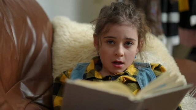 A preteen girl reading a book begins to yawn, while lying on the sofa in the living room of a modern house