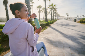 View from the back to a smiling athlete woman drinking water after morning jog on the promenade