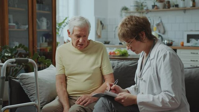 Female doctor in white coat sitting on couch, asking elderly man about his health and filling out medical record form during consultation at home
