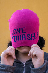 Girl wearing pink cap with self love message and pulling it, yellow background
