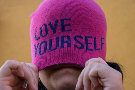 Girl wearing pink cap with self love message and pulling it, yellow background