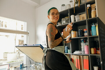 Positive female worker standing with bank of ink on background of own design studio and looks camera