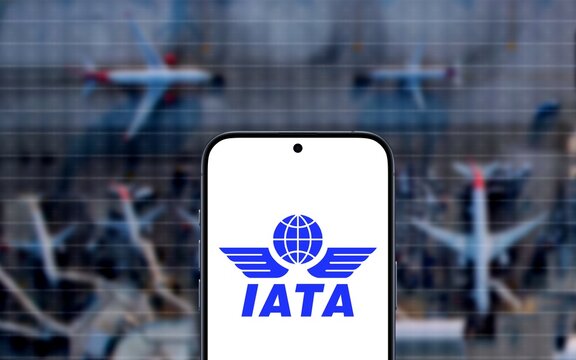 IATA Logo is shown on a modern smartphone, International Air Transport Association, in the background an airport with airplanes and world map with connections, aviation, regulations