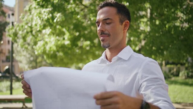 Smiling draftsman examining plan at sunlight city closeup. Man holding blueprint