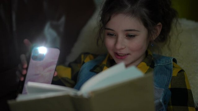 Close-up. Happy preteen girl reading a book using a flashlight from her mobile phone, lying in the dark on the sofa in the living room of a modern house