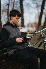 A contemplative young adult relaxes with a book on a park bench, with his bicycle parked nearby, embodying leisure and tranquility.