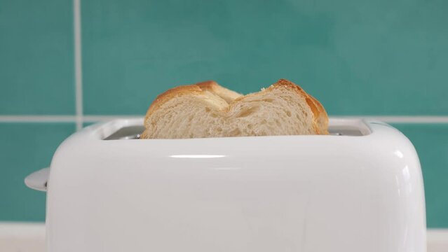 Piece of bread pops out of a white toaster against background of mint tiles in the kitchen.