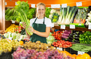 Portrait of positive teenage girl in uniform working in grocery shop as job experience, selling celery