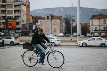 A woman rides a bike in the city, wearing a casual outfit and beanie, with distant mountains and...