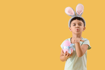 Cute little boy wearing bunny ears with Easter gift egg blowing kiss on yellow background