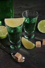 Absinthe in shot glasses, spoon, brown sugar cubes and lime on gray textured table, closeup....