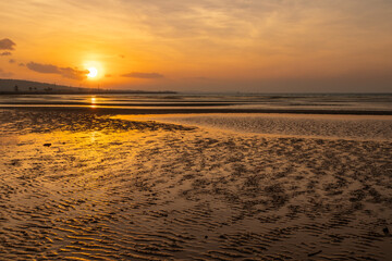 A sunrise through clouds over a bay at low tide showing sand ripples in the reflections at the town...