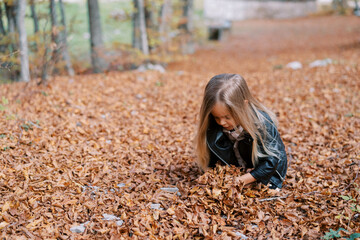 Little girl squats, raking dry fallen leaves into a pile with her hands in the autumn forest