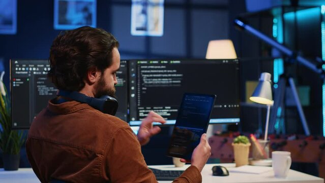 Software developer programming in home office, crosschecking lines of code between tablet and PC screen. IT admin holding portable device, using it as reference while coding on computer, camera A