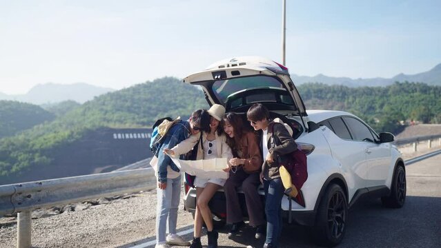 Group of Happy Asian man and woman enjoy and fun outdoor lifestyle road trip travel nature countryside on summer holiday vacation. Generation z people friends sitting on car trunk and looking on a map