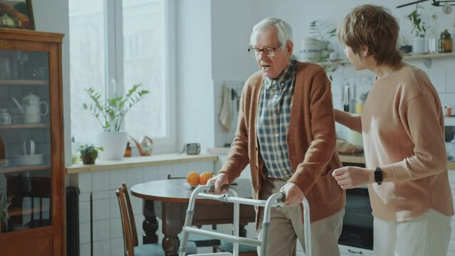 Elderly man using walking frame at home with help of adult granddaughter walking beside and telling him supportive words