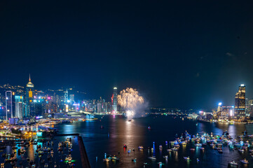 Fototapeta na wymiar Fireworks burst brightly against the night sky above a city skyline, reflecting in the calm water below
