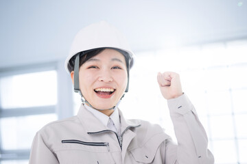 メインビジュアルにも！逆光の中作業着を着たカメラ目線のヘルメットを被る女性　ガッツポーズでやる気のイメージのクローズアップ