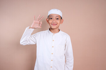 Little Asian muslim boy with skullcap showing ok sign with finger over beige background