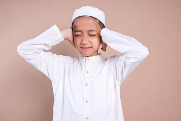 Young muslim boy wear skullcap covering his ears with two hands, distressed by noise isolated over...