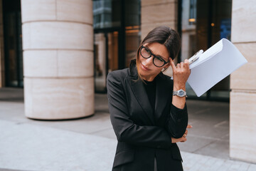 Confident businesswoman in glasses with documents outside modern building, looking puzzled