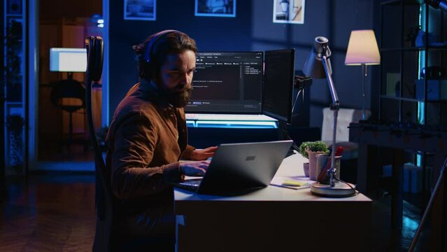 Developer focused on typing complex code in neon lit home office, listening music and developing software application. IT expert wearing headphones, solving coding project tasks on laptop, camera B