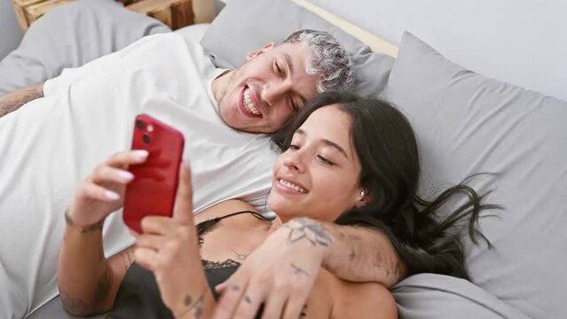 A cheerful man and woman sharing a joyful moment with a smartphone in a cozy bedroom