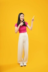 The photo of a young Asian woman with a satisfied expression, standing and pointing away with both forefingers, wearing a pink t-shirt, isolated on a yellow background. Advertisement concept.