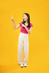 The photo of a young Asian woman with a satisfied expression, standing and pointing away with both forefingers, wearing a pink t-shirt, isolated on a yellow background. Advertisement concept.