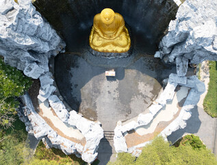 Aerial view of golden Buddha statue with waterfall and stone wall in background at Wat Lak Si Rat Samoson, Samut Sakhon, Thailand.