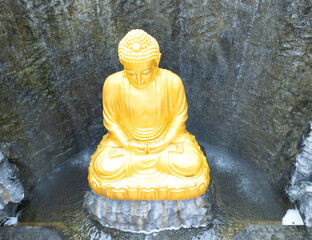 Top view of golden Buddha statue with waterfall and stone wall in background at Wat Lak Si Rat Samoson, Samut Sakhon, Thailand.