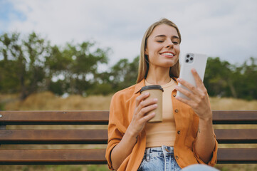 Young happy student fun woman wears orange shirt casual clothes use mobile cell phone drink coffee sit on bench walk rest relax in spring green city park outdoors on nature. Urban lifestyle concept.