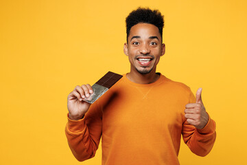 Young man wear orange sweatshirt casual clothes hold eat bar of chocolate show thumb up gesture...