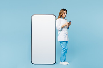 Full body doctor woman in white medical gown suit work in hospital clinic office big huge blank screen mobile cell phone use smartphone isolated on plain blue background Health care medicine concept