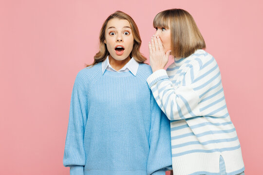 Elder parent mom 50s year old with young adult daughter two women together in blue casual clothes whisper gossip and tells secret behind her hand isolated on plain pink background Family day concept