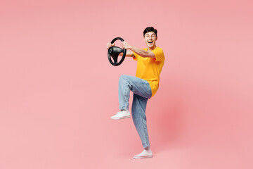Full body sideways young man he wearing yellow t-shirt casual clothes holding steering wheel driving car raise up leg isolated on plain pastel light pink background studio portrait. Lifestyle concept.