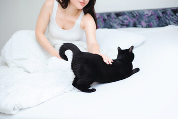 Young beautiful woman in a white tank top petting her black cat on her white bed - 742356802