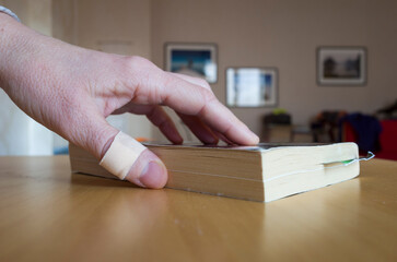 A hand with an adhesive bandage on the thumb takes a book from a wooden table on a blurred...