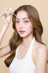 Young Asian beauty woman curly long hair with korean makeup style on face and using pipette to apply serum on skin on isolated beige background. Facial treatment, Cosmetology, plastic surgery.