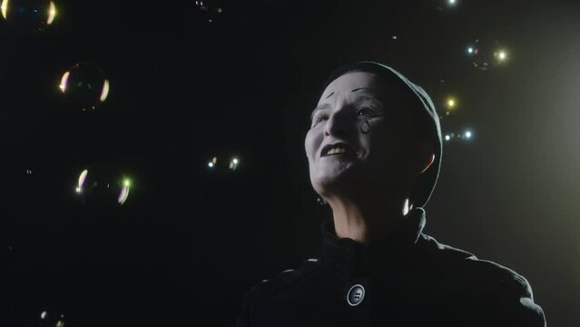 Chest up slowmo of rejoiced mime woman looking around at flying soap bubbles in black background