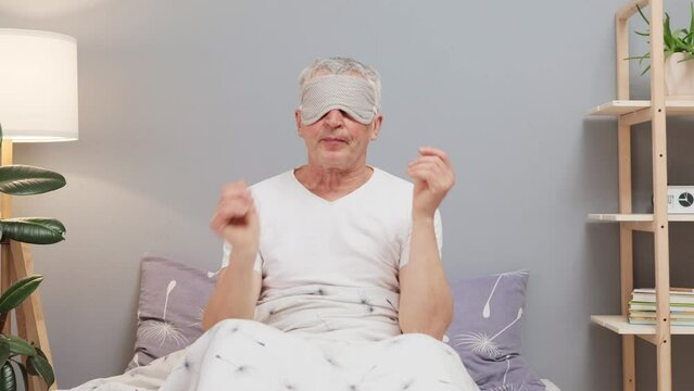 Sleepy senior man waking up in the morning in his bed at home having sleepless night suffering insomnia tsking off sleeping mask having tired unhappy facial expression