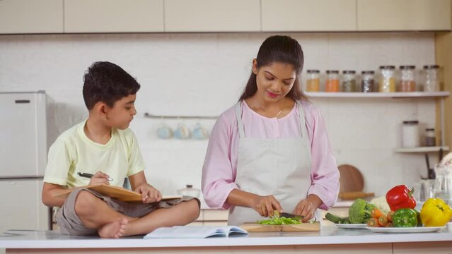 mother multitasking by helping child with homework while chopping vegetables at kitchen - concept of motherhood, responsibility and education assistance
