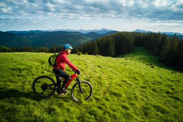 Obraz premium Cyclist man riding electric bike outdoors on sunny evening. Male tourist resting on grassy hill, enjoying beautiful mountain landscape, wearing helmet and backpack. Concept of active leisure.