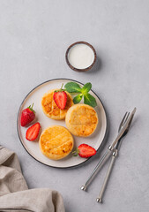 Cottage cheese, cheesecakes or syrniki with strawberries and mint leaves on a white plate on a light background with sour cream and cutlery.