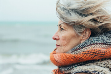 Thoughtful senior woman with a cold, wearing a scarf, stands by the sea, gazing into the distance - 742442279