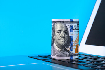 Laptop with a roll of paper one hundred dollar bills on a blue background. Copy space.