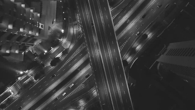 Black and white footage of drone top down aerial of cars drive at of bridge and underpass at night with streaks from moving cars and motorbikes. Complex markings on main road.