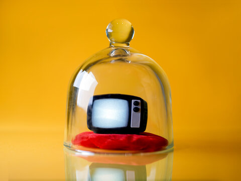 Mini tv on a red pillow under a glass cloche against a yellow background
